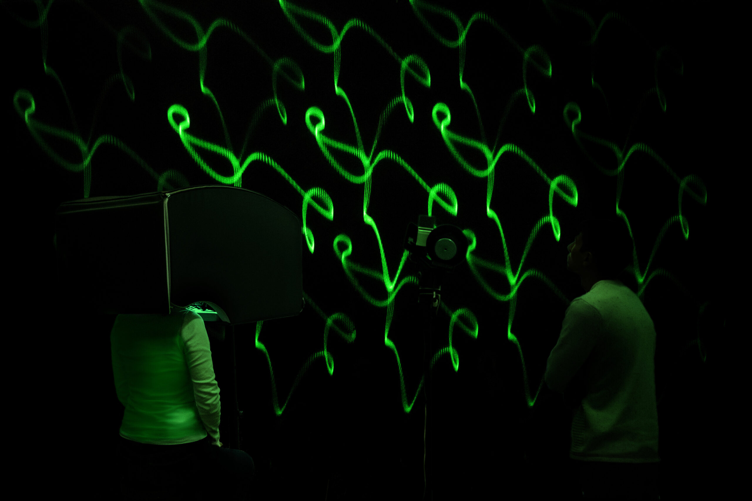 Two people stand in front of a wall on which green lasers make twisting shapes. The person on the left stands with their head inside of a console. The person on the right looks at the wall.