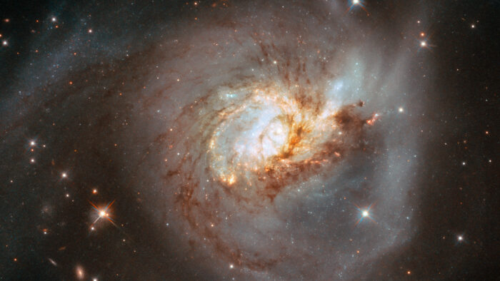 A large, face-on spiral galaxy. The core is radiating very brightly in white and bluish colours. Thin webs of dark gas cross the centre and most of the galaxy, following the twist of the galaxy. This gas continues out into a tidal tail at the upper-left, and another at the bottom. Small stars can be seen around the spiral galaxy, on a black background